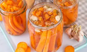 Gläser mit karotten fermentieren, gefüllt mit Karottenstücken, Knoblauch und Kräutern. Daneben liegen frische Karottenscheiben und eine Knoblauchknolle.