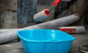 Kleister mit idealer kleister konsistenz tropft vom Malerroller in eine blaue Wanne. Tapetenrollen liegen bereit.