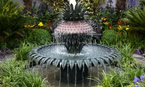 Ein kunstvoller Gartenbrunnen im Ananas-Design, der zum **gartenbrunnen selber bauen** inspiriert. Umgeben von üppiger Vegetation und bunten Blumen.