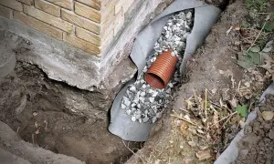 Drainage verlegen: Ein orangefarbenes Rohr liegt in einer Kiesschicht, umgeben von Vlies und Erdreich neben einer Ziegelwand.