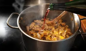 Rotwein wird beim Kochen in einen Topf mit Fleisch und Zwiebeln gegossen. Dampf steigt auf, während das Gericht köchelt.