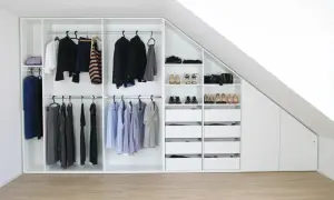 Ein maßgeschneiderter schrank selber bauen: weiße Kleiderschränke mit Kleiderstangen, Schubladen und Regalen, gefüllt mit Kleidung und Schuhen, unter einer Dachschräge.