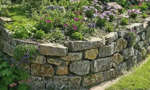 Eine **trockenmauer** sichert den **Hang** mit Natursteinen. Blumen und Grünpflanzen wachsen üppig darauf und daneben.