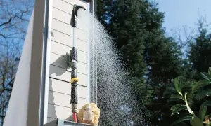 Gartendusche selber bauen: Eine Dusche mit Schlauchanschluss und Brausekopf sprüht Wasser vor einer Holzwand.