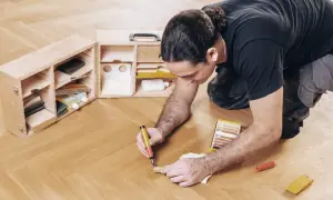 Mann repariert Parkett mit einem Reparaturstift und einer Farbkarte. Werkzeugkasten mit Zubehör daneben.