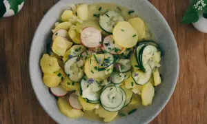 Ein Teller mit bayrischem Kartoffelsalat mit Gurke, Radieschen und Schnittlauch, garniert mit kleinen blauen Blüten.
