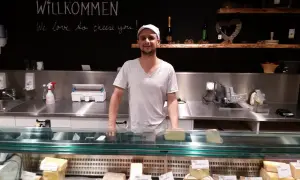 Ein Mann steht hinter einer Käsetheke. Die Tafel über ihm verkündet "Willkommen, we love to cheese you!". Hier gibt es nur das Beste, auch mit tierischem Lab.