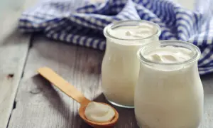 Zwei Gläser cremigen griechischen Joghurts, reich an Protein, stehen auf einem Holztisch. Ein Holzlöffel mit einer Portion Joghurt liegt daneben.
