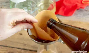Flüssigkeit wird aus einer braunen Flasche durch einen Kaffeefilter in ein Glas gegossen. So gelingt essig selber machen.