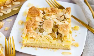 Ein Stück veganer Stachelbeerkuchen mit Streuseln und Mandelblättchen auf einem weißen Teller, daneben goldene Gabeln.
