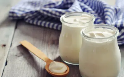Zwei Gläser cremigen griechischen Joghurts, reich an Protein, stehen auf einem Holztisch. Ein Holzlöffel mit einer Portion Joghurt liegt daneben.