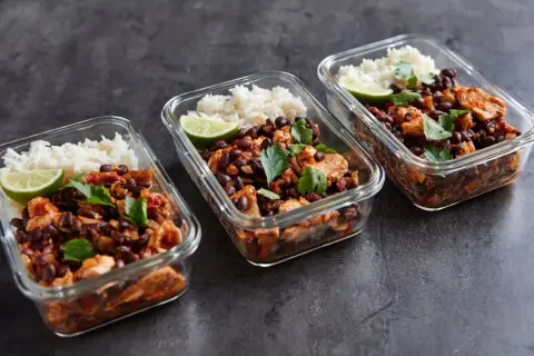 Drei Glasbehälter mit meal prep reis, Hähnchen, schwarzen Bohnen und Limettenspalten, garniert mit Petersilie.