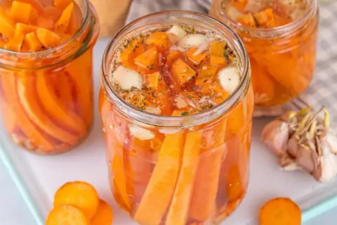 Gläser mit karotten fermentieren, gefüllt mit Karottenstücken, Knoblauch und Kräutern. Daneben liegen frische Karottenscheiben und eine Knoblauchknolle.