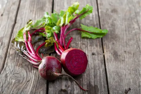 Zwei frische rote Bete, eine davon halbiert, liegen auf rustikalem Holz. Perfekt, um rote Beete roh zu essen und die gesunden Nährstoffe zu genießen.