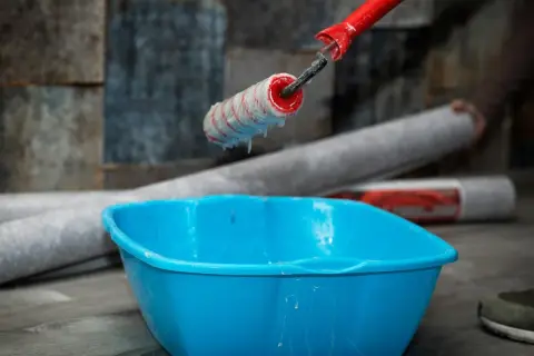 Kleister mit idealer kleister konsistenz tropft vom Malerroller in eine blaue Wanne. Tapetenrollen liegen bereit.