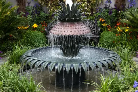Ein kunstvoller Gartenbrunnen im Ananas-Design, der zum **gartenbrunnen selber bauen** inspiriert. Umgeben von üppiger Vegetation und bunten Blumen.
