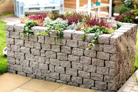 Ein bepflanztes Hochbeet mit stabilen hochbeet mauern aus Betonsteinen, umgeben von grünem Rasen und einer Terrasse.