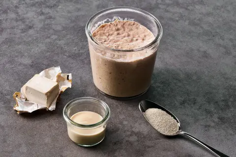 Frischer Hefewürfel, ein Glas mit Sauerteig, eine kleine Schale mit Milch und ein Löffel mit Trockenhefe – Zutaten für einen Hefe Ersatz.