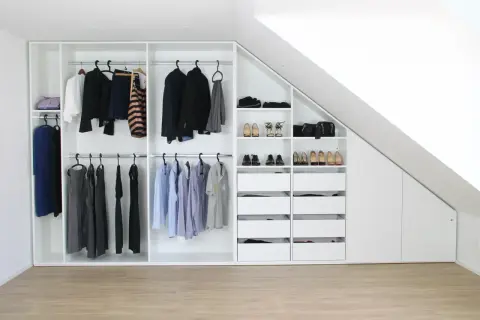 Ein maßgeschneiderter schrank selber bauen: weiße Kleiderschränke mit Kleiderstangen, Schubladen und Regalen, gefüllt mit Kleidung und Schuhen, unter einer Dachschräge.