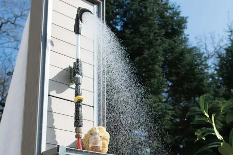 Gartendusche selber bauen: Eine Dusche mit Schlauchanschluss und Brausekopf sprüht Wasser vor einer Holzwand.