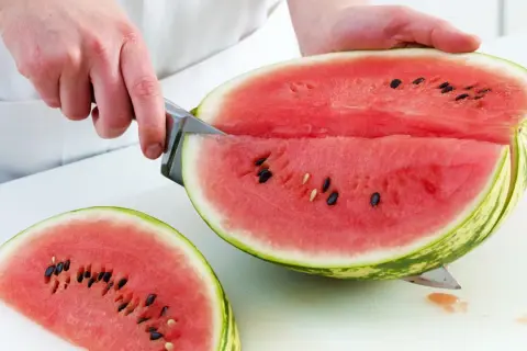 So schneidest du eine Wassermelone richtig: Ein Messer teilt die saftige Frucht in zwei Hälften, eine davon liegt bereits als Stück daneben.