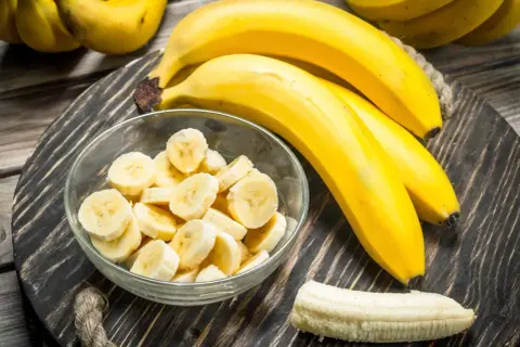 Bananen auf einem Holzteller, eine Schüssel mit Bananenscheiben und eine geschälte Banane. Interessiert, wie viel Zucker hat eine Banane?