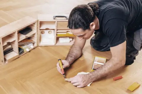 Mann repariert Parkett mit einem Reparaturstift und einer Farbkarte. Werkzeugkasten mit Zubehör daneben.