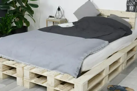Ein rustikales Bett, das man einfach selber bauen kann, aus Europaletten. Mit grauer Decke und Kissen.