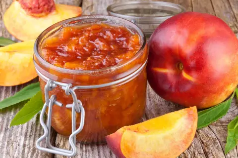 Glas mit leuchtend orangefarbener Nektarinen Marmelade, daneben frische Nektarinen und Fruchtstücke auf rustikalem Holz.