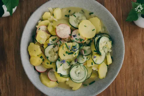 Ein Teller mit bayrischem Kartoffelsalat mit Gurke, Radieschen und Schnittlauch, garniert mit kleinen blauen Blüten.