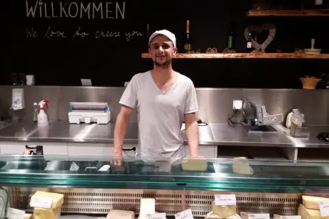 Ein Mann steht hinter einer Käsetheke. Die Tafel über ihm verkündet "Willkommen, we love to cheese you!". Hier gibt es nur das Beste, auch mit tierischem Lab.