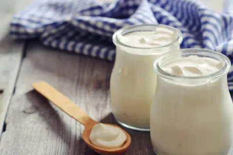 Zwei Gläser cremigen griechischen Joghurts, reich an Protein, stehen auf einem Holztisch. Ein Holzlöffel mit einer Portion Joghurt liegt daneben.