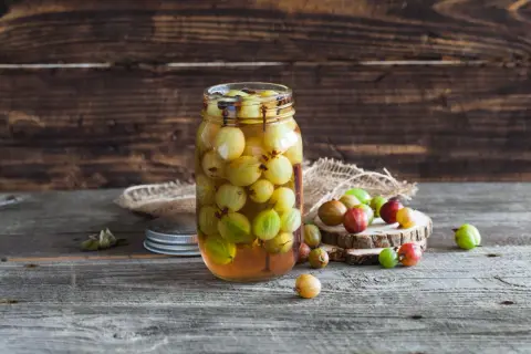 Ein Glas mit stachelbeeren einkochen, daneben liegen einige frische Beeren auf Holzscheiben.