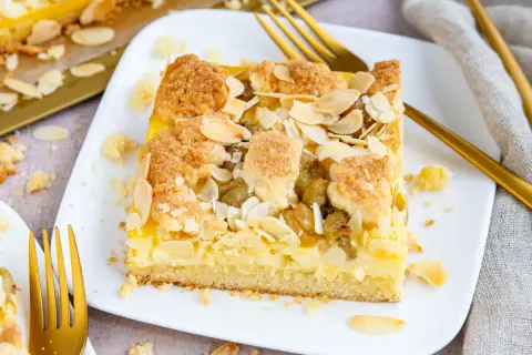 Ein Stück veganer Stachelbeerkuchen mit Streuseln und Mandelblättchen auf einem weißen Teller, daneben goldene Gabeln.
