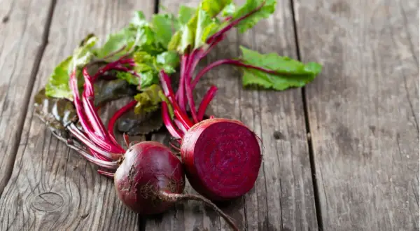 Zwei frische rote Bete, eine davon halbiert, liegen auf rustikalem Holz. Perfekt, um rote Beete roh zu essen und die gesunden Nährstoffe zu genießen.