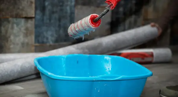 Kleister mit idealer kleister konsistenz tropft vom Malerroller in eine blaue Wanne. Tapetenrollen liegen bereit.