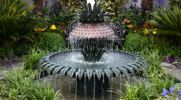 Ein kunstvoller Gartenbrunnen im Ananas-Design, der zum **gartenbrunnen selber bauen** inspiriert. Umgeben von üppiger Vegetation und bunten Blumen.