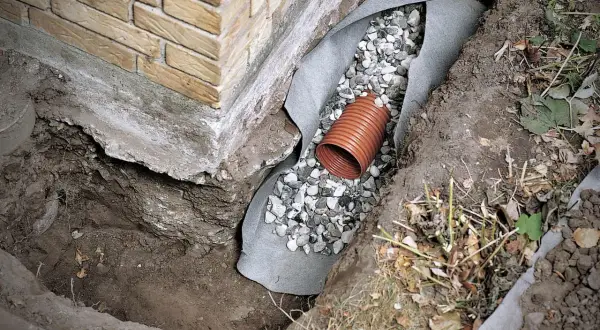 Drainage verlegen: Ein orangefarbenes Rohr liegt in einer Kiesschicht, umgeben von Vlies und Erdreich neben einer Ziegelwand.