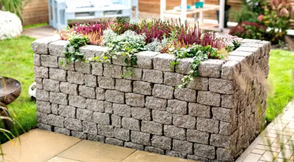 Ein bepflanztes Hochbeet mit stabilen hochbeet mauern aus Betonsteinen, umgeben von grünem Rasen und einer Terrasse.