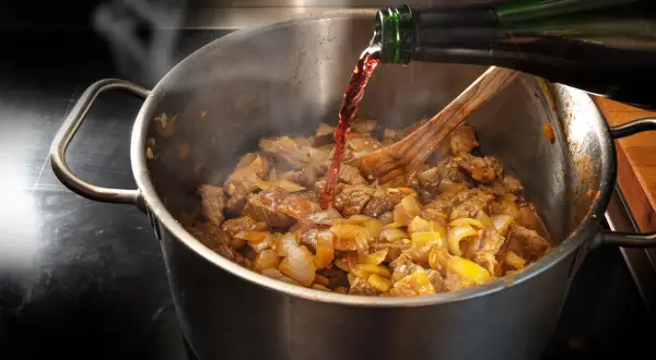 Rotwein wird beim Kochen in einen Topf mit Fleisch und Zwiebeln gegossen. Dampf steigt auf, während das Gericht köchelt.