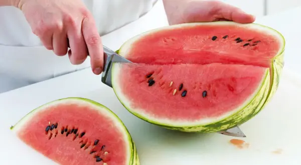 So schneidest du eine Wassermelone richtig: Ein Messer teilt die saftige Frucht in zwei Hälften, eine davon liegt bereits als Stück daneben.