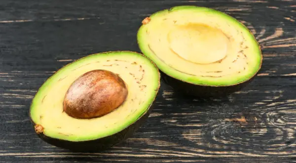 Eine aufgeschnittene Avocado mit Kern und einigen kleinen schwarzen Punkten im Fruchtfleisch liegt auf dunklem Holz.