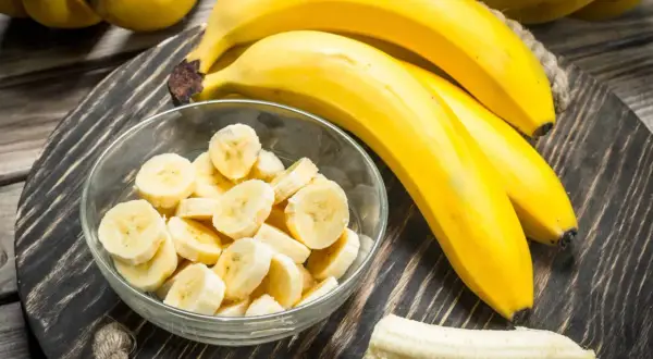 Bananen auf einem Holzteller, eine Schüssel mit Bananenscheiben und eine geschälte Banane. Interessiert, wie viel Zucker hat eine Banane?