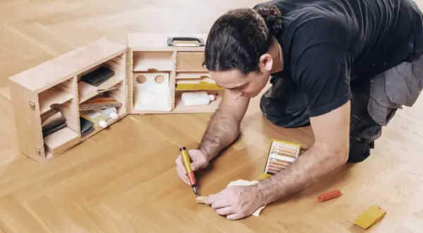 Mann repariert Parkett mit einem Reparaturstift und einer Farbkarte. Werkzeugkasten mit Zubehör daneben.
