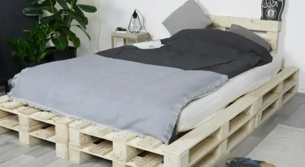 Ein rustikales Bett, das man einfach selber bauen kann, aus Europaletten. Mit grauer Decke und Kissen.