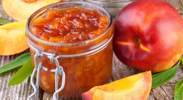 Glas mit leuchtend orangefarbener Nektarinen Marmelade, daneben frische Nektarinen und Fruchtstücke auf rustikalem Holz.