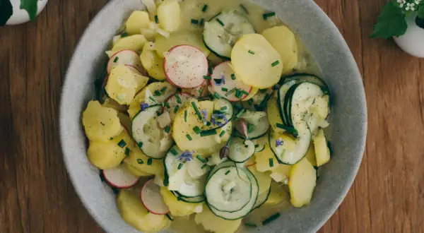 Ein Teller mit bayrischem Kartoffelsalat mit Gurke, Radieschen und Schnittlauch, garniert mit kleinen blauen Blüten.