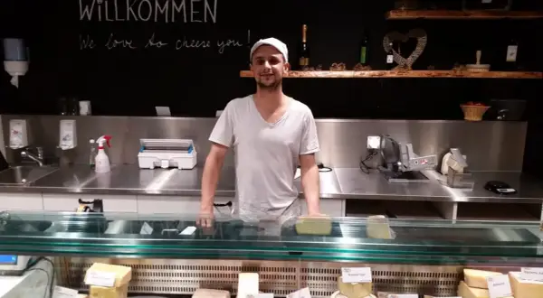 Ein Mann steht hinter einer Käsetheke. Die Tafel über ihm verkündet "Willkommen, we love to cheese you!". Hier gibt es nur das Beste, auch mit tierischem Lab.