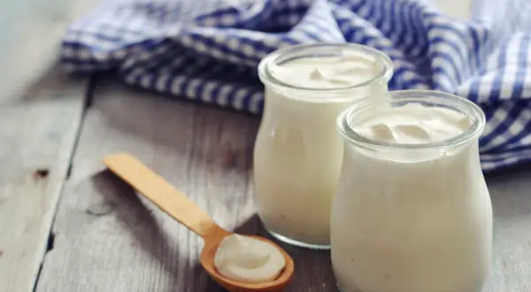 Zwei Gläser cremigen griechischen Joghurts, reich an Protein, stehen auf einem Holztisch. Ein Holzlöffel mit einer Portion Joghurt liegt daneben.