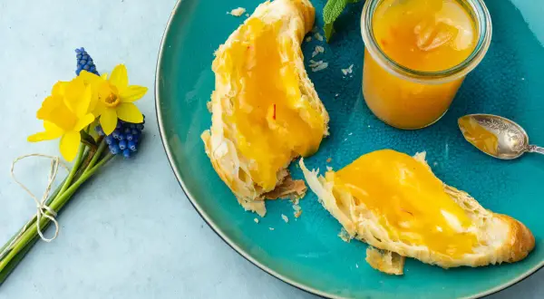 Quittenrezepte Marmelade auf Brot, daneben ein Glas mit der leuchtend gelben Marmelade und ein Löffel. Ein Frühlingsstrauß mit Narzissen und Traubenhyazinthen.